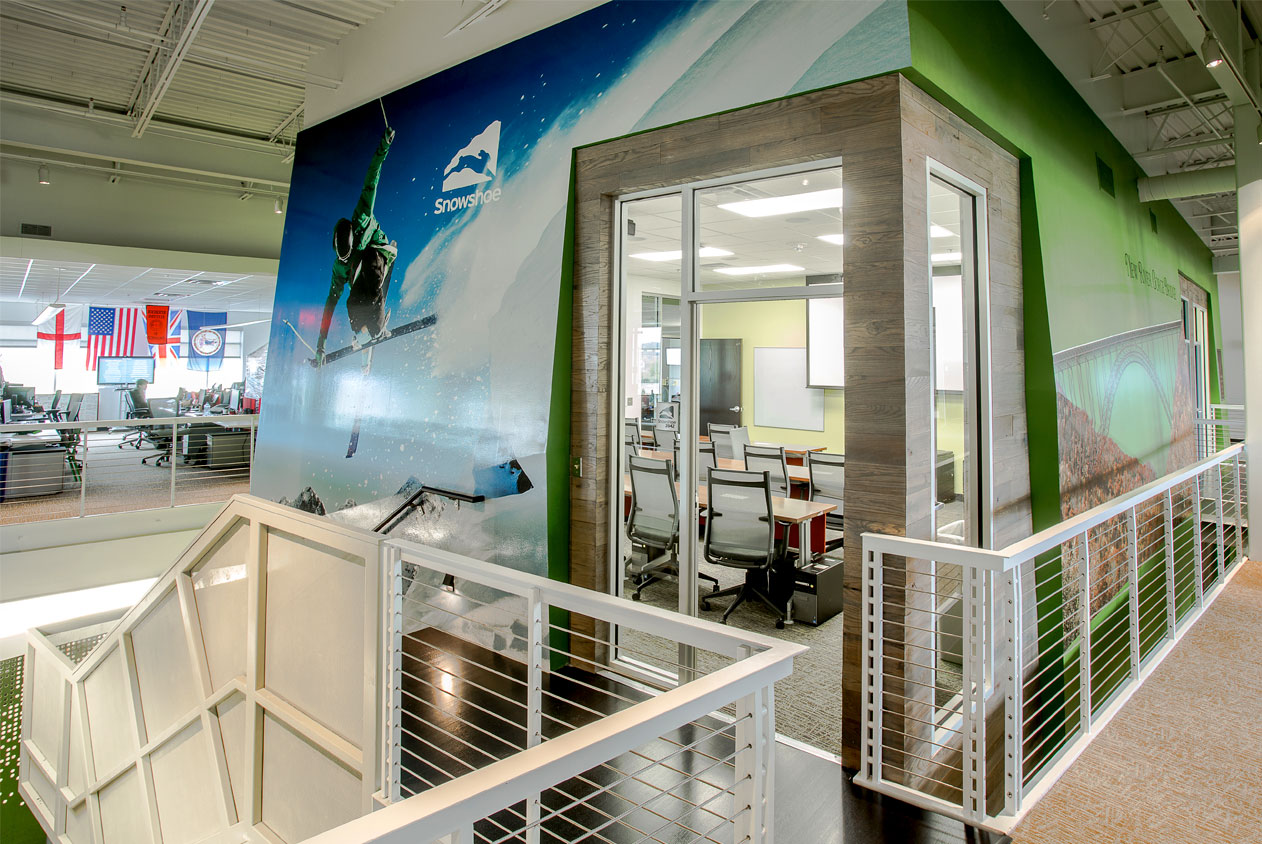 Rackspace Office Design: Virginia - A view from a staircase landing overlooking an office space. A large mural of a skier jumping a ramp dominates one wall. A glass-walled meeting room is visible in the background.