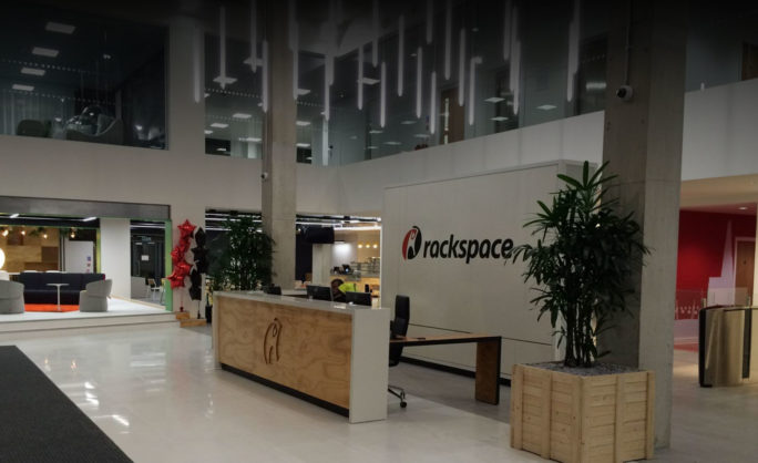 Rackspace Office Design: London - Modern Rackspace office lobby featuring a minimalist design with a wooden reception desk, hanging linear lights, indoor plants, and a red-accented waiting area