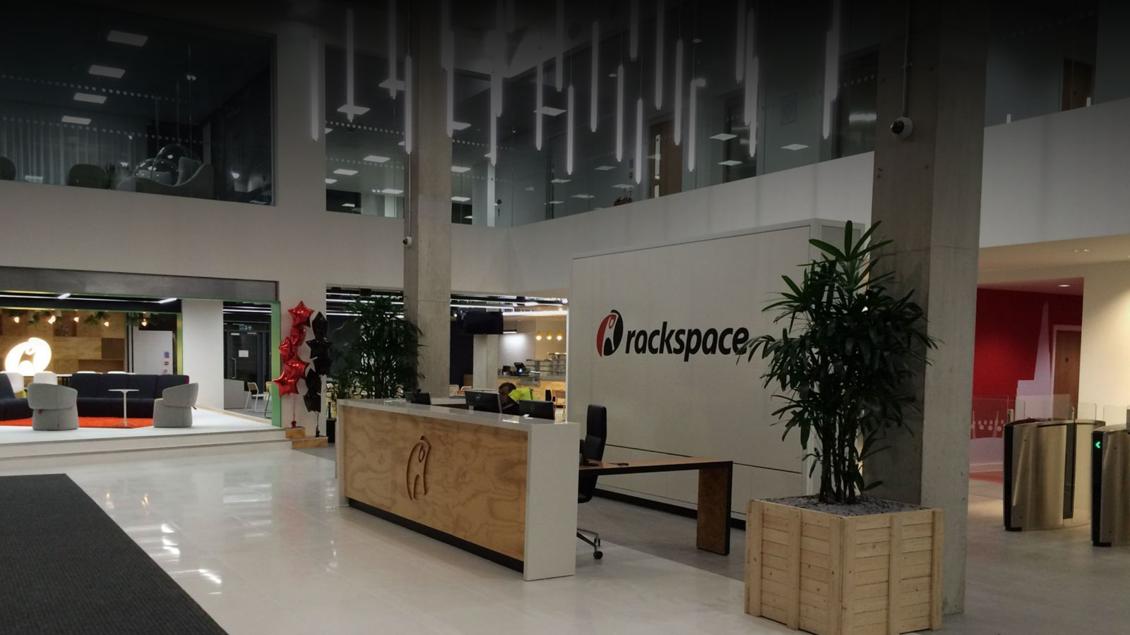 Rackspace Office Design: London - Modern Rackspace office lobby featuring a minimalist design with a wooden reception desk, hanging linear lights, indoor plants, and a red-accented waiting area