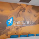 A photo of a conference room with a large mural on the wall. The mural features a close-up image of a child building with blocks, emphasizing creativity and learning. The VentureLab logo, featuring a rocket ship, is prominently displayed alongside the text "Entrepreneurship Education for Kids."