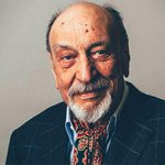 A portrait of Milton Glaser, a renowned graphic designer. He is shown with a warm smile, wearing a dark suit and a colorful scarf.