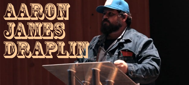 Aaron James Draplin delivering a presentation at a podium, wearing a blue cap and denim jacket, with bold typography featuring his name