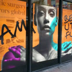 Storefront window display featuring a bold design with a grayscale portrait of a woman, colorful accents in teal and purple, and large black text stating ‘I AM BEAUTIFUL’ over an orange background with faint text.