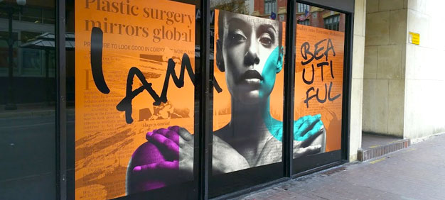 Storefront window display featuring a bold design with a grayscale portrait of a woman, colorful accents in teal and purple, and large black text stating ‘I AM BEAUTIFUL’ over an orange background with faint text.