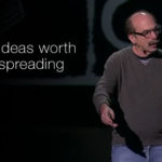 Speaker presenting on stage at a TED talk, with the TED logo and tagline ‘Ideas worth spreading’ displayed