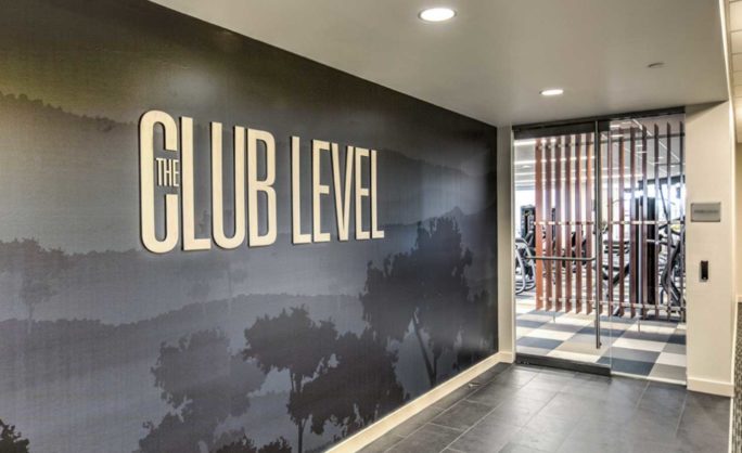 The Club Level - Entrance to ‘The Club Level’ featuring a sleek, modern design with a shadowed tree mural on the wall and a view into a gym through glass doors