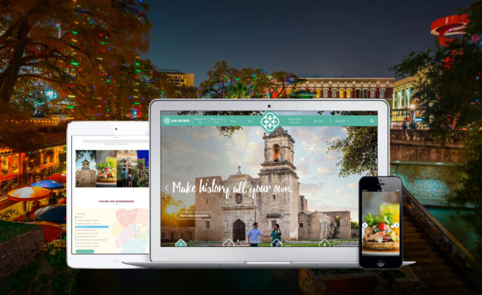 Visit San Antonio - Visit San Antonio website displayed on various devices including a laptop, tablet, and smartphone, set against a festive nighttime Riverwalk scene illuminated with colorful lights