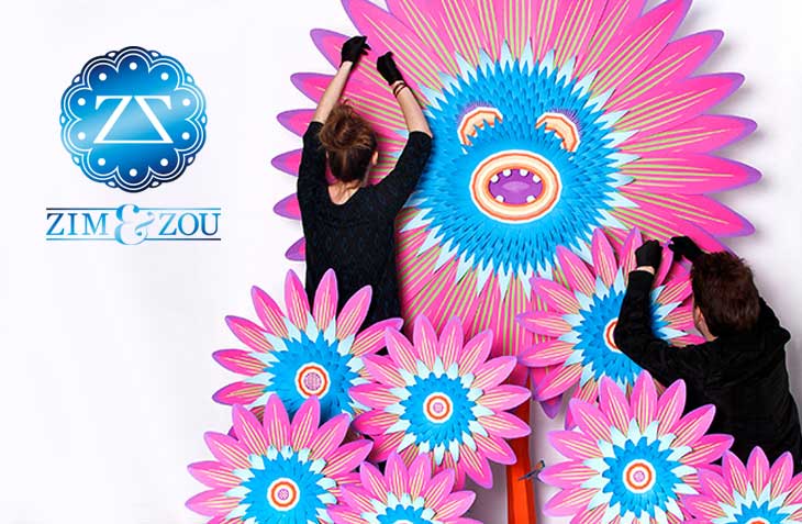 Artists Zim and Zou assembling vibrant pink, blue, and white paper art floral designs with a whimsical face centerpiece.