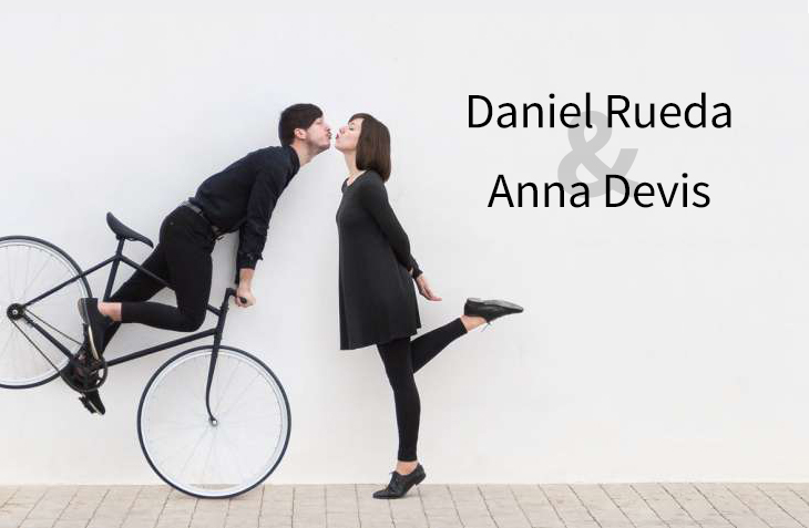 Daniel Rueda and Anna Devis creatively posing with a bicycle against a minimalist white background.