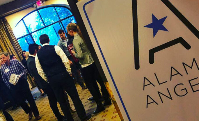 A networking event featuring attendees engaged in conversations near a large sign displaying the Alamo Angels logo, set against a room with golden curtains and natural light from arched windows.