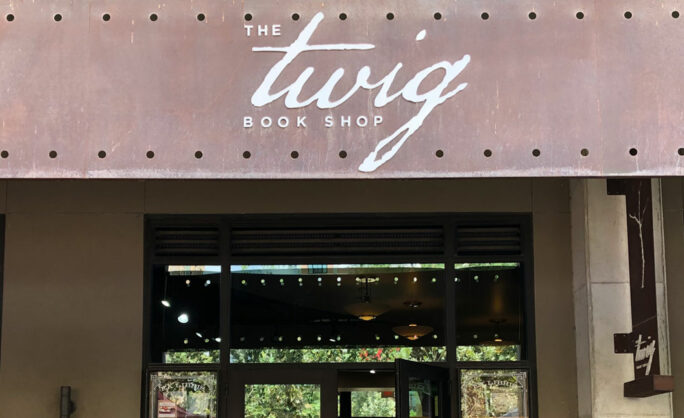 The front entrance of The Twig Book Shop, showcasing its logo in a stylish handwritten font on a rusted, perforated metal panel. The store's glass doors and windows allow a glimpse into the warm interior ambiance